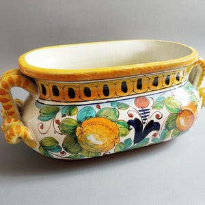 May include: Oval ceramic planter with a cream interior and a yellow rim. The exterior features hand-painted floral and fruit designs in orange, green, and blue. It has two ornate yellow handles.