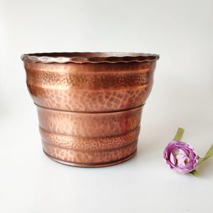 Może przedstawiać: Doniczka z młotkowanego miedzi z falistym brzegiem. Doniczka jest jasno miedziana i ma lekko zużyty wygląd.