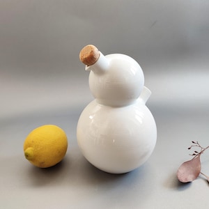 Könnte beinhalten: Eine weiße Ölflasche aus Keramik mit einem Korken. Links von der Flasche liegt eine gelbe Zitrone. Die Flasche hat eine runde Form mit einem Ausguss. Rechts befindet sich ein Zweig mit getrockneten Blättern.