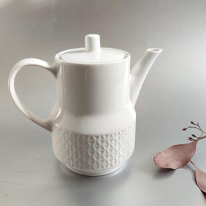 May include: White ceramic teapot with a lid and a handle. The lower portion of the teapot features a geometric pattern. The teapot is designed with a spout for pouring. A small sprig of dried leaves is in the background.