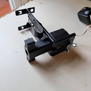 May include: A black plastic 3D printed bracket with a metal clip and a small black motor attached to it. The bracket is designed to hold a device or object.