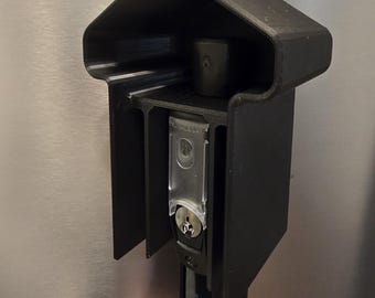 May include: Black plastic gate latch with a key lock and a protective hood. The latch has a rectangular design with a clear plastic cover over the lock mechanism. The text "MAGNALatch" is visible on the lower part of the latch.