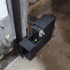 May include: A black garage door opener sensor with a green light. The sensor is mounted to a metal door frame.