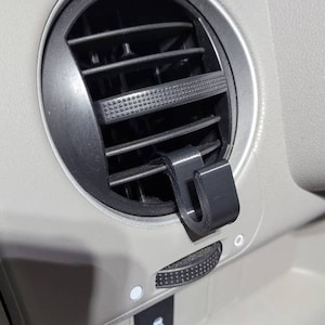 May include: A close-up of a car's air vent with a black plastic hook attached to the vent. The hook is designed to hold items like grocery bags or other small objects.