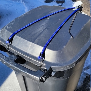 Puede incluir: Un contenedor de basura negro con un cord&oacute;n el&aacute;stico azul que asegura la tapa. El contenedor est&aacute; sobre una superficie de hormig&oacute;n con nieve en el fondo.