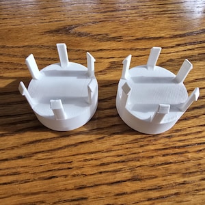 Op de afbeelding: Twee witte, cirkelvormige, 3D-geprinte objecten met verticale steunen aan de rand. Ze liggen op een houten oppervlak. De steunen zijn gelijkmatig verdeeld over de omtrek van de cirkelvormige basis. Elk object heeft een diameter van ongeveer 7 cm.