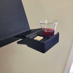 May include: A black plastic holder with a small white square and a clear cup filled with red liquid. The holder is attached to a black surface.