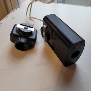 May include: A black flash unit with a small black remote trigger. The flash unit has a textured surface and a round hole on the side. The remote trigger has a small green light and a screw mount.