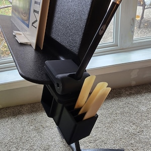 Puede incluir: Un atril negro con un libro, un lápiz y cañas. El atril tiene una bandeja para partituras, un soporte para lápices y un compartimento para cañas. El atril está sobre una base de trípode negra.