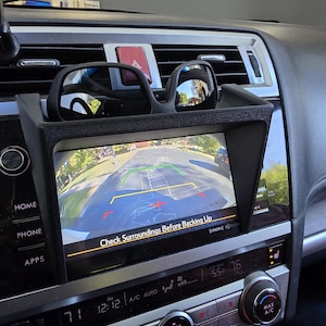 Puede incluir: Un salpicadero de coche con una pantalla integrada que muestra la vista de la cámara de marcha atrás. Gafas de sol negras descansan sobre la pantalla. El texto "Check Surroundings Before Backing Up" se muestra. La temperatura es de 24 grados Celsius.