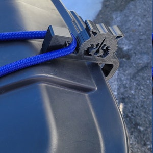 Puede incluir: Un soporte de tapa de cubo de basura de pl&aacute;stico negro con una cuerda azul atada. El soporte tiene forma de cabeza de lobo.