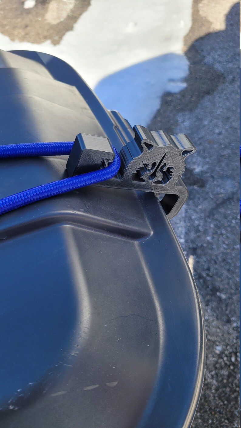 Puede incluir: Un soporte de tapa de cubo de basura de pl&aacute;stico negro con una cuerda azul atada. El soporte tiene forma de cabeza de lobo.