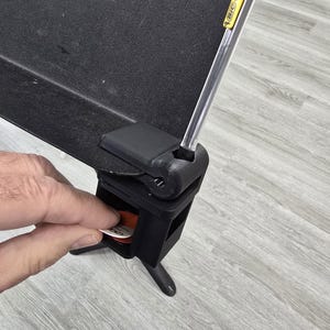May include: Black adjustable music stand with a built-in storage compartment. A yellow and clear BIC pencil rests on the stand. A hand is reaching into the compartment to retrieve a guitar pick. The stand is on a gray wooden floor.