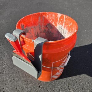 Op de afbeelding: Een oranje emmer met grijze en rode gereedschappen bevestigd. De emmer is gevuld met een grijze substantie. Een grijze gereedschapshouder is aan de zijkant van de emmer bevestigd. De emmer staat op een donkergrijze ondergrond.