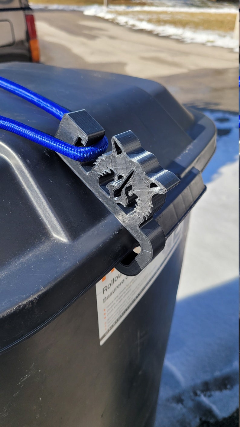 Puede incluir: Un contenedor de basura negro con un clip de pl&aacute;stico gris sujeto a la parte superior. El clip tiene un dise&ntilde;o de lobo y una cuerda azul est&aacute; unida a &eacute;l. El texto "Rollout Basurero" es visible en el lateral del contenedor.
