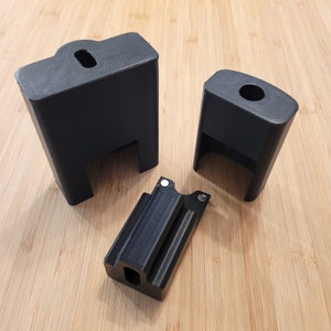 May include: Three black plastic 3D printed parts, two are rectangular with a hole in the top and one is a smaller rectangular piece with a slot on one side.