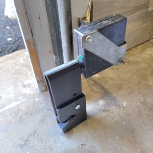 May include: A black metal garage door sensor with a rectangular black box and a silver metal bracket. The sensor is mounted on a metal arm and has a green light.
