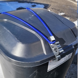Puede incluir: Un contenedor de basura negro con un cord&oacute;n el&aacute;stico azul que asegura la tapa. El contenedor tiene una etiqueta que dice "Rollout  Basureros".
