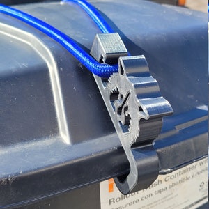 Puede incluir: Un contenedor de basura de pl&aacute;stico negro con un cord&oacute;n el&aacute;stico azul sujeto a un gancho de pl&aacute;stico negro. El contenedor de basura tiene una etiqueta que dice "Rollout Trash Container with Basurero con tapa abatible y ruedas".