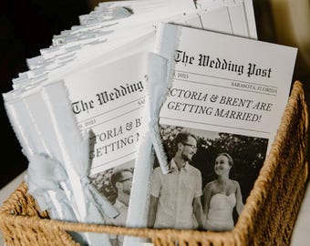 May include: A wicker basket holds stacks of wedding newspapers. Each paper is headed "The Wedding Post" and displays a photo of a couple with the headline "Victoria & Brent Are Getting Married!" Lit candles in glass holders are visible.