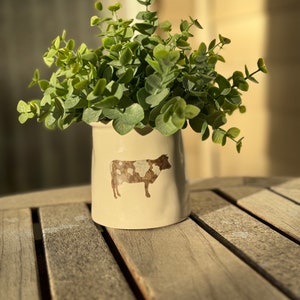 May include: A cream-colored ceramic vase with a cow illustration, filled with green leafy plants. The vase sits on a wooden table, with sunlight casting shadows. The cow illustration is brown and white.