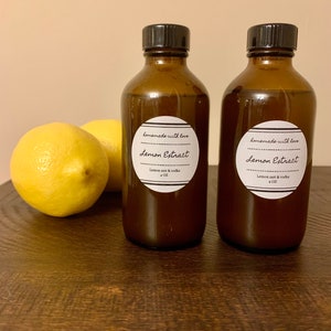 Könnte beinhalten: Zwei bernsteinfarbene Glasflaschen mit schwarzen Deckeln, gefüllt mit einer dunklen Flüssigkeit. Jede Flasche hat ein weißes Etikett mit schwarzem Text, auf dem "homemade with love Lemon Extract Lemon zest & vodka 4 OZ" steht.