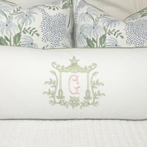 May include: A long, white decorative pillow with a green border and an embroidered design. The design features a pagoda-style structure with the letter "G" in pink. Two floral patterned pillows are in the background.
