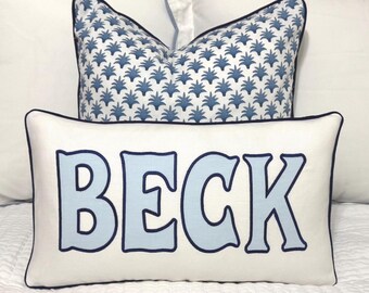 May include: White pillow with blue trim and the name "BECK" in light blue letters. The pillow is on top of a white pillow with a blue and white patterned pillow behind it.