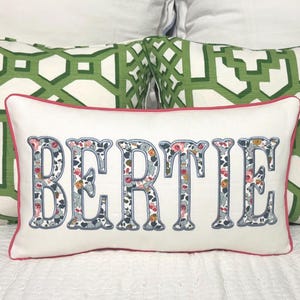 Op de afbeelding: Wit rechthoekig kussen met de naam "BERTIE" in letters met bloemenpatroon, blauwe omlijning en roze rand. Twee groen-wit patronen kussens op de achtergrond.