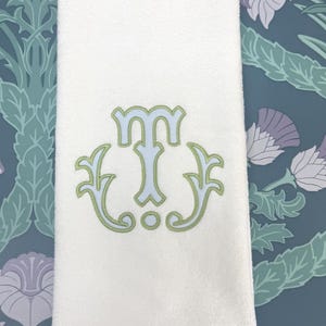 Op de afbeelding: Witte badhanddoek met een geborduurd monogram in lichtblauw en groen. Het monogram is een sierlijk ontwerp, met een gestileerde letter "M" in het midden. De handdoek heeft een getextureerd oppervlak.