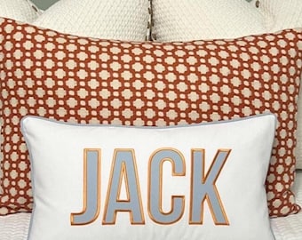 May include: White pillow with the name "JACK" embroidered in blue and gold letters. The pillow is on top of a red and white patterned pillow.