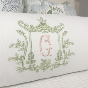May include: White rectangular pillow with a green embroidered pagoda design and the letter "G" in pink. The pillow has a green trim and is placed on a white surface. Decorative pillow for a bed or sofa.