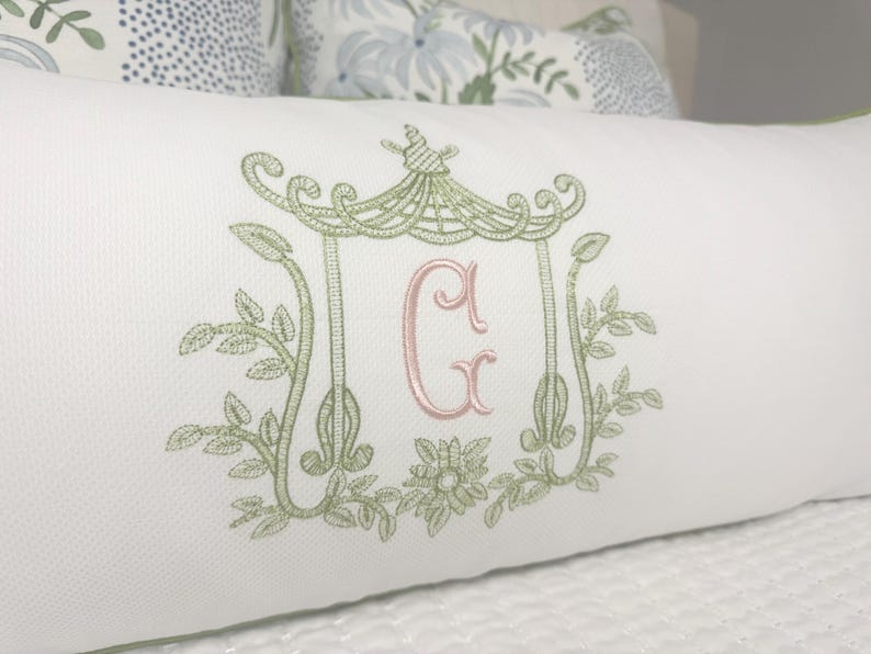 May include: White rectangular pillow with a green embroidered pagoda design and the letter "G" in pink. The pillow has a green trim and is placed on a white surface. Decorative pillow for a bed or sofa.