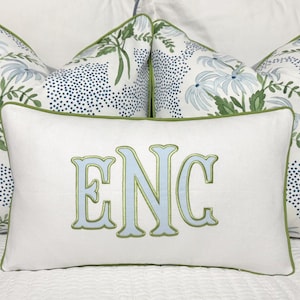 May include: A set of decorative pillows with a white rectangular pillow in front, embroidered with the letters "ENC" in light blue and outlined in green. The background pillows have a floral pattern with blue and green accents.
