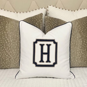 Op de afbeelding: Wit kussen met een marineblauwe rand en een gouden en marineblauwe monogram van de letter "H" in een vierkant frame.