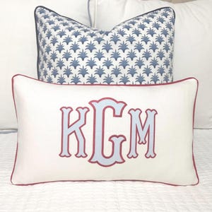 May include: Two decorative pillows on a bed. The front pillow is white with a red border and light blue embroidered initials KGM. The back pillow has a blue and white floral pattern. The pillows are on a white bedspread.