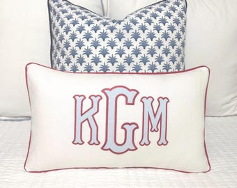 May include: Two decorative pillows on a bed. The front pillow is white with a red border and light blue embroidered initials KGM. The back pillow has a blue and white floral pattern. The pillows are on a white bedspread.