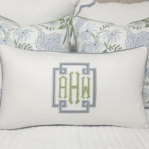Op de afbeelding: Wit rechthoekig kussen met een monogram in een grijs en groen geometrisch ontwerp. Twee kussens met een bloemenpatroon in blauw, groen en wit staan erachter. De kussens liggen op een wit sprei.