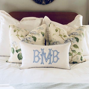 May include: A white bed with a white and green floral patterned pillow, a white pillow with blue embroidery that says "BMB", and a white duvet cover.