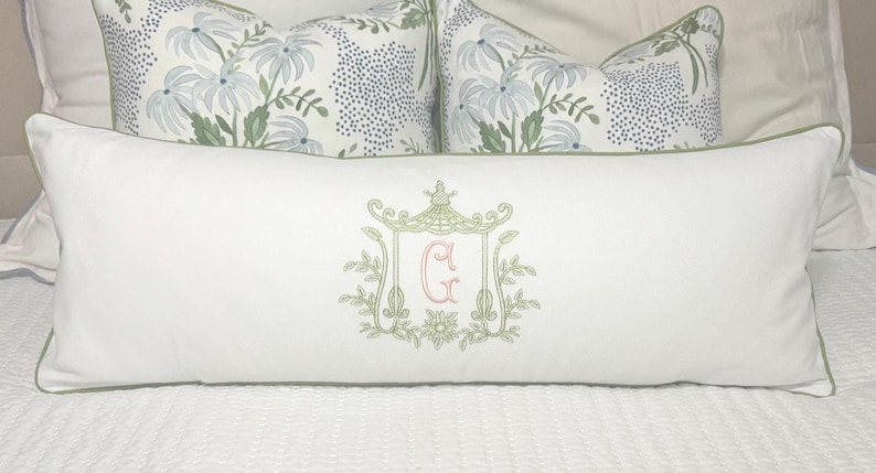 May include: A long, white decorative pillow with a green border and an embroidered design. The design features a pagoda-style structure with the letter "G" in pink. Two floral patterned pillows are in the background.