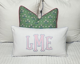 May include: White pillow with a light blue and pink monogram "LME" embroidered on the front. The pillow has a light blue trim and is sitting on a white bed with a green and white patterned pillow behind it.