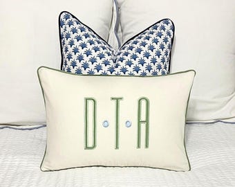 May include: A white pillow with a green border and the monogram "D.T.A." embroidered in green. The pillow is sitting on a white bed with a blue and white patterned pillow behind it.
