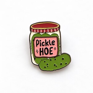 May include: A metal enamel pin shaped like a glass jar filled with green pickles. The jar features a pink label with the black text Pickle HOE and two small star icons. The lid is red with vertical stripes, and a single pickle rests in front.