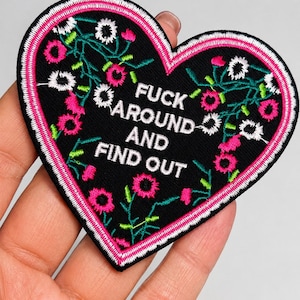 May include: A heart-shaped embroidered patch with a black background and a pink and white border. The patch features white text that reads FUCK AROUND AND FIND OUT, surrounded by embroidered floral vines in shades of pink, white, and green.