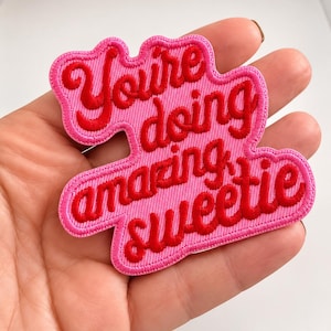 May include: An embroidered patch with a pink background and red cursive text that reads "You're doing amazing sweetie". The patch has a scalloped, irregular shape that follows the contour of the lettering, featuring a dense thread texture.