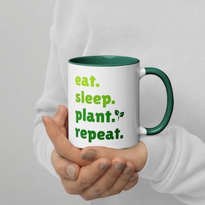 以下が含まれることがあります： 緑色の縁と取っ手のある白いセラミック製のマグカップ。マグカップには緑色の文字で「eat. sleep. plant. repeat.」と書かれています