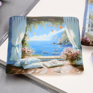 May include: Square decorative tile with a painted coastal scene. The image depicts a view through an open window with blue ocean, boats, and a mountain in the distance. White curtains frame the view, with flowers and pillows in the foreground.