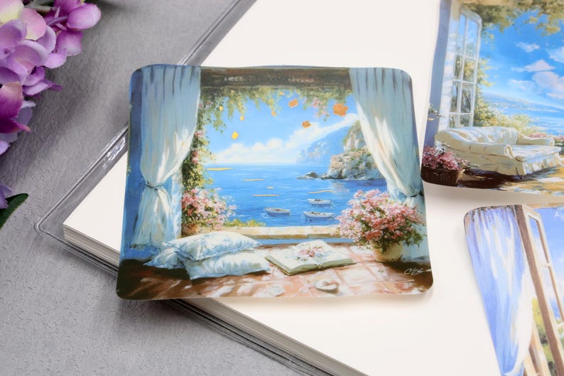 May include: Square decorative tile with a painted coastal scene. The image depicts a view through an open window with blue ocean, boats, and a mountain in the distance. White curtains frame the view, with flowers and pillows in the foreground.