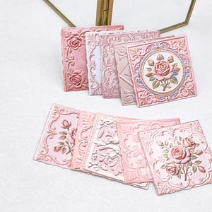 Op de afbeelding: Een verzameling vierkante tegels met reliëf bloemmotieven in roze en witte tinten. De tegels tonen ingewikkelde reliëfontwerpen van rozen, bladeren en sierlijke krulranden, waarbij sommige stukken een centraal rozenmotief bevatten.
