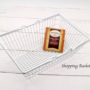 May include: White wire shopping basket with a single tea bag inside. The tea bag is labeled "Twinings Earl Grey Black Tea". The text "Shopping Basket L" is visible below the basket.